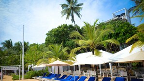 Eine Strandlounge mit weißen Liegestühlen und Sonnenschirmen, einem Schild mit der Aufschrift "KINGFISHER" und üppiger Bepflanzung im Hintergrund.