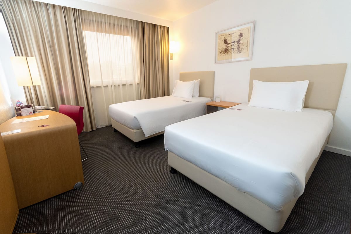 Habitación de hotel con dos camas, un escritorio, una silla y un ventanal con cortinas.