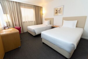 A hotel room with two beds, a desk, a chair, and a large window with curtains.