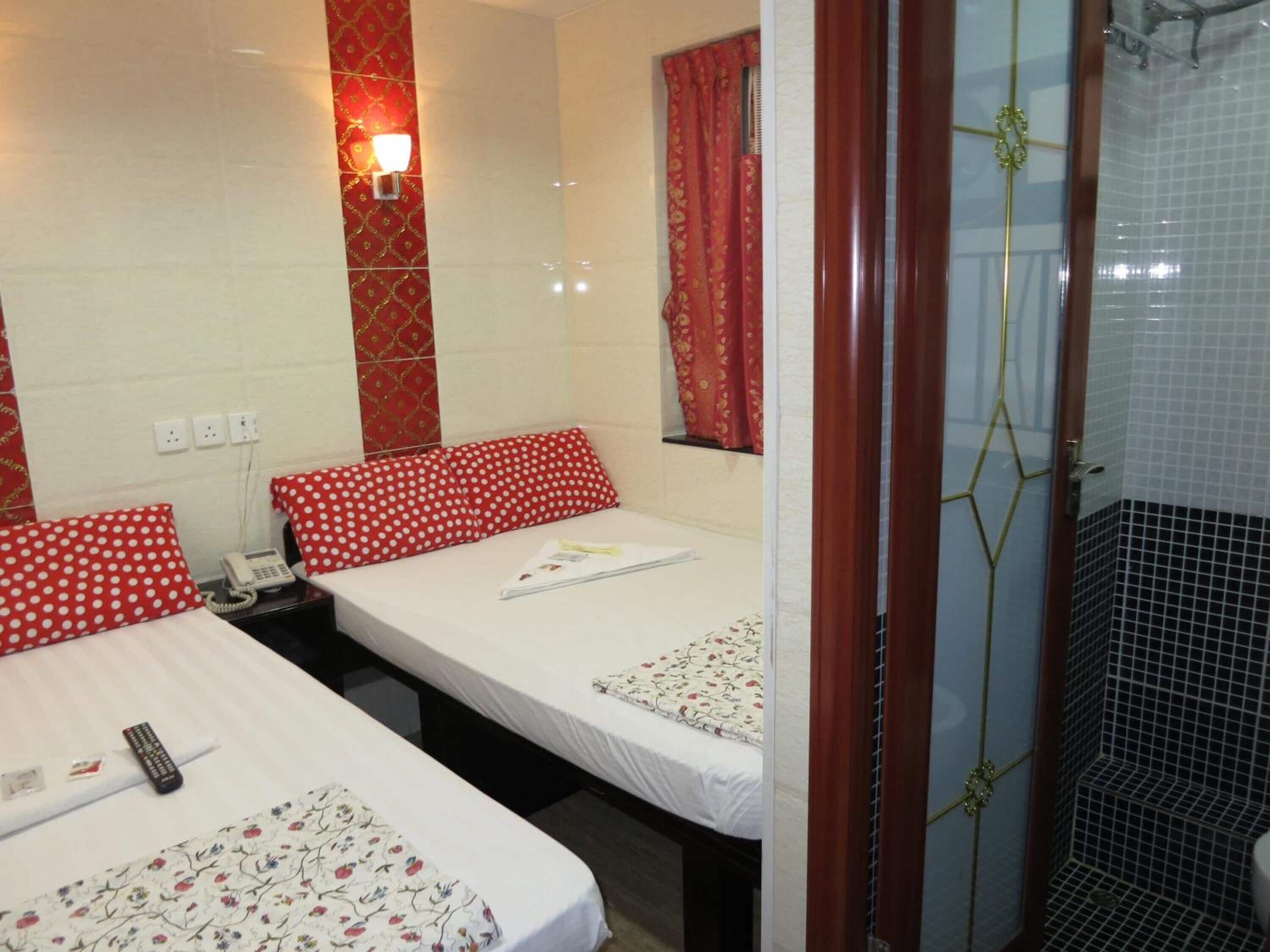 Habitación de hotel con dos camas, cada una con un cojín rojo de lunares y una colcha con estampado floral. Se aprecia una mesita de noche con un teléfono y un control remoto. Se ve una ventana con cortinas rojas, y una zona de ducha con puerta de vidrio al fondo.