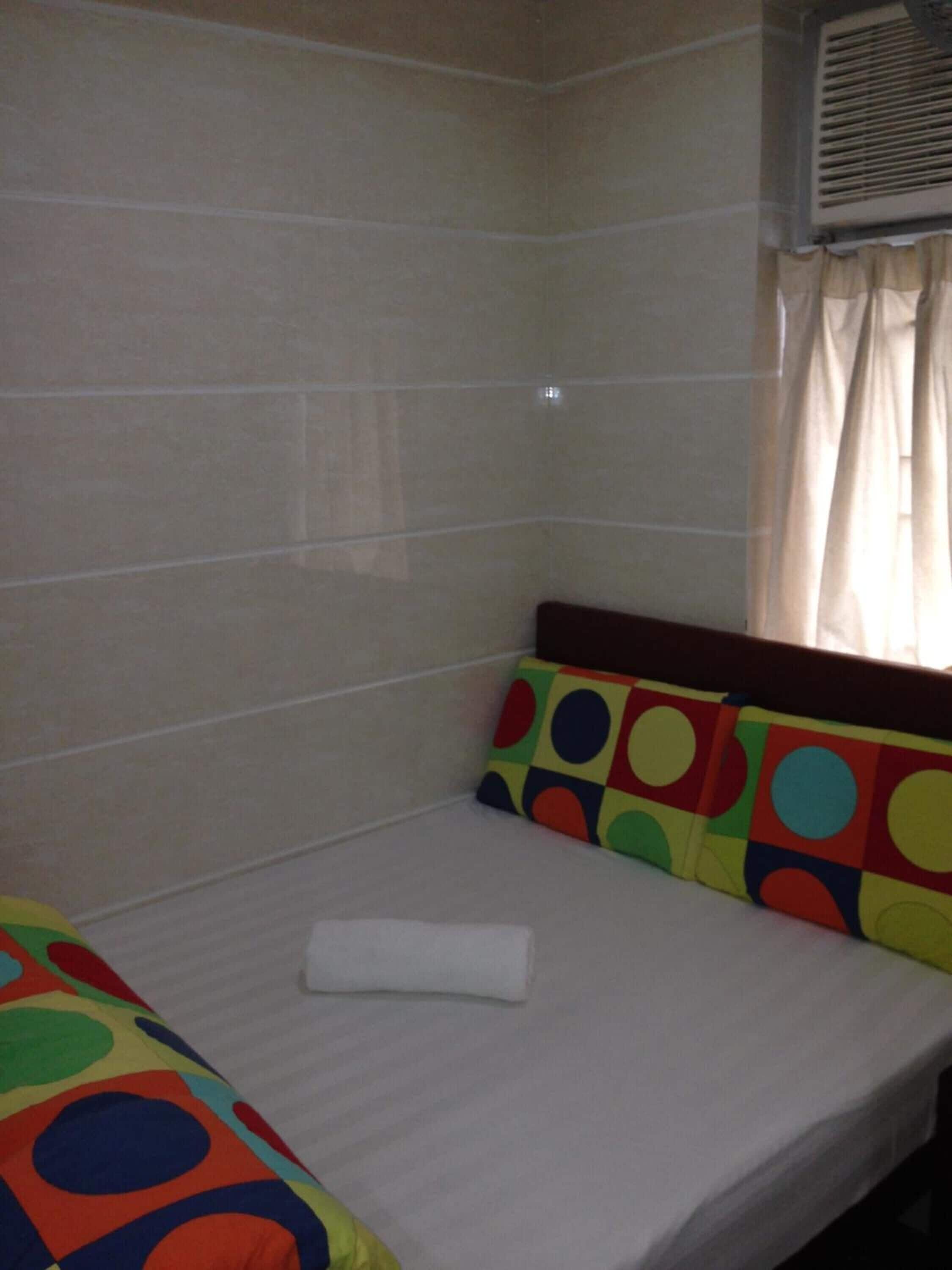 A small, single-bed room with a tiled wall, a window with curtains, and colorful pillows.