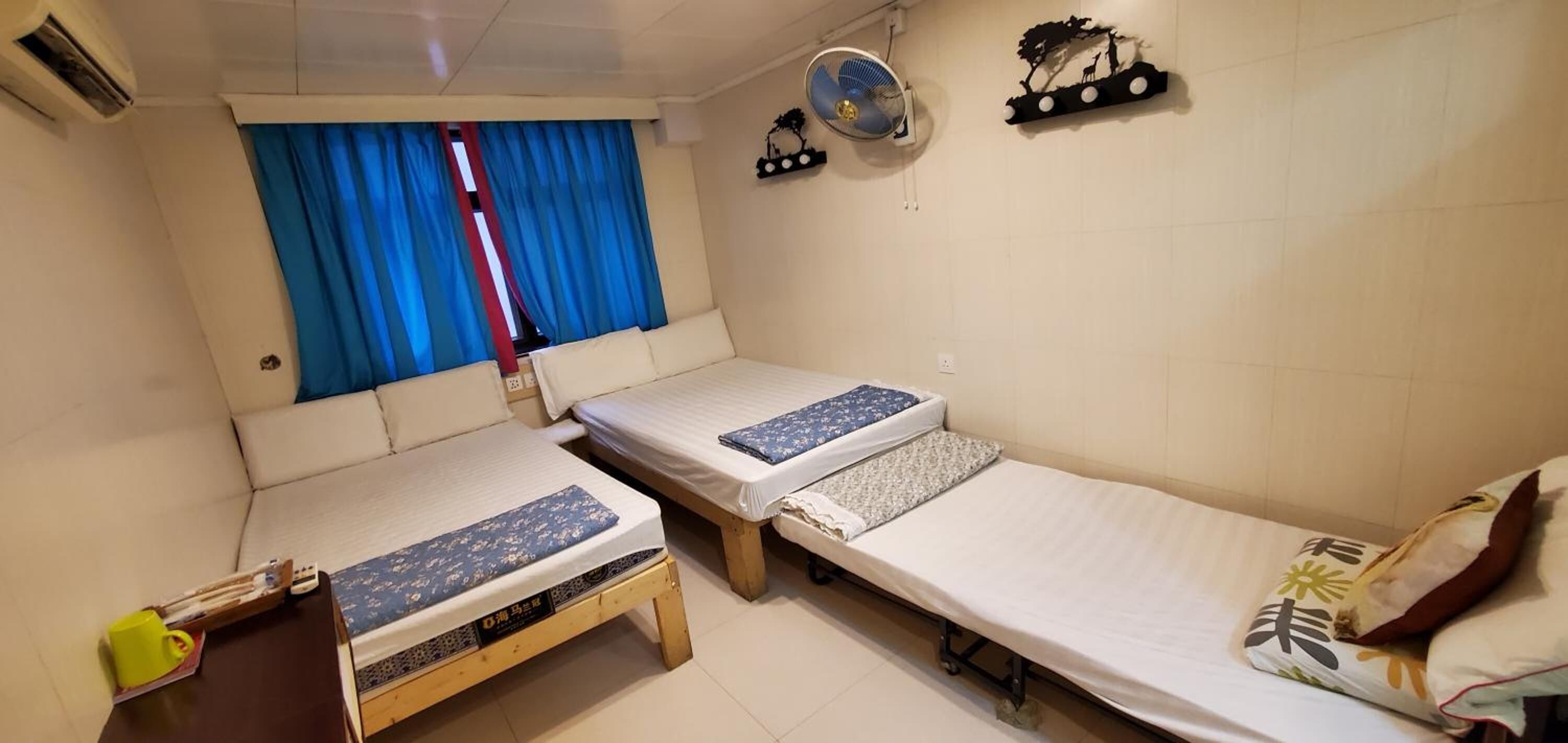 Une petite chambre propre, avec deux lits simples, une table de chevet, un ventilateur et une fenêtre avec des rideaux bleus.