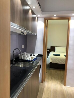 A compact kitchen with a sink, stove, and cabinets, leading to a small bedroom with a bed and nightstand. - Cebu Hostel (Kowloon)