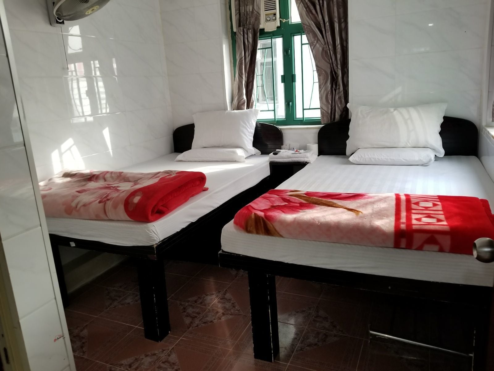 Zwei Einzelbetten mit weißen Laken und roten Decken, ein Fenster mit Vorhängen und ein gefliester Boden.