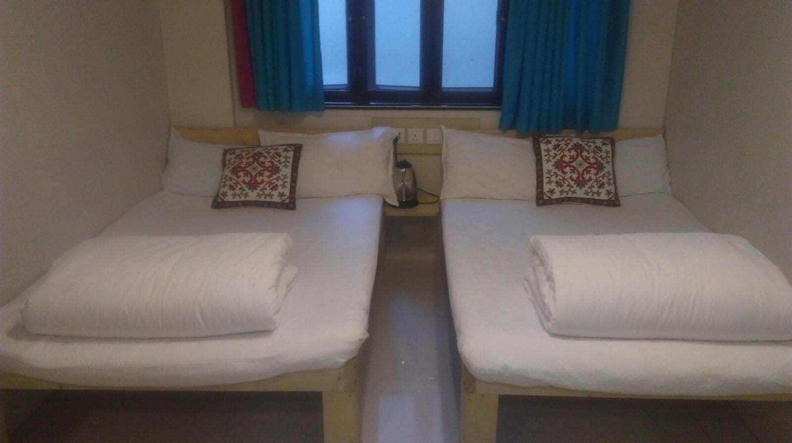 Una habitación con dos camas individuales, cada una con almohada y manta. Hay una ventana con cortinas, una mesita de noche con lámpara y un enchufe de pared.