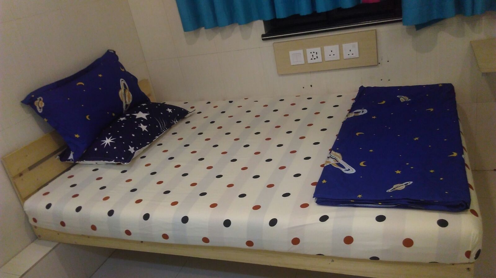 A small, single-bed room with a polka dot bedspread, two pillows with space-themed designs, and a window with blue curtains.