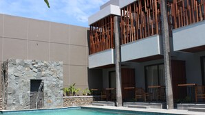 A pool area with a stone wall, wooden deck, and a modern building with balconies. - Grand Laguna Hotel & Villa (Colomadu)
