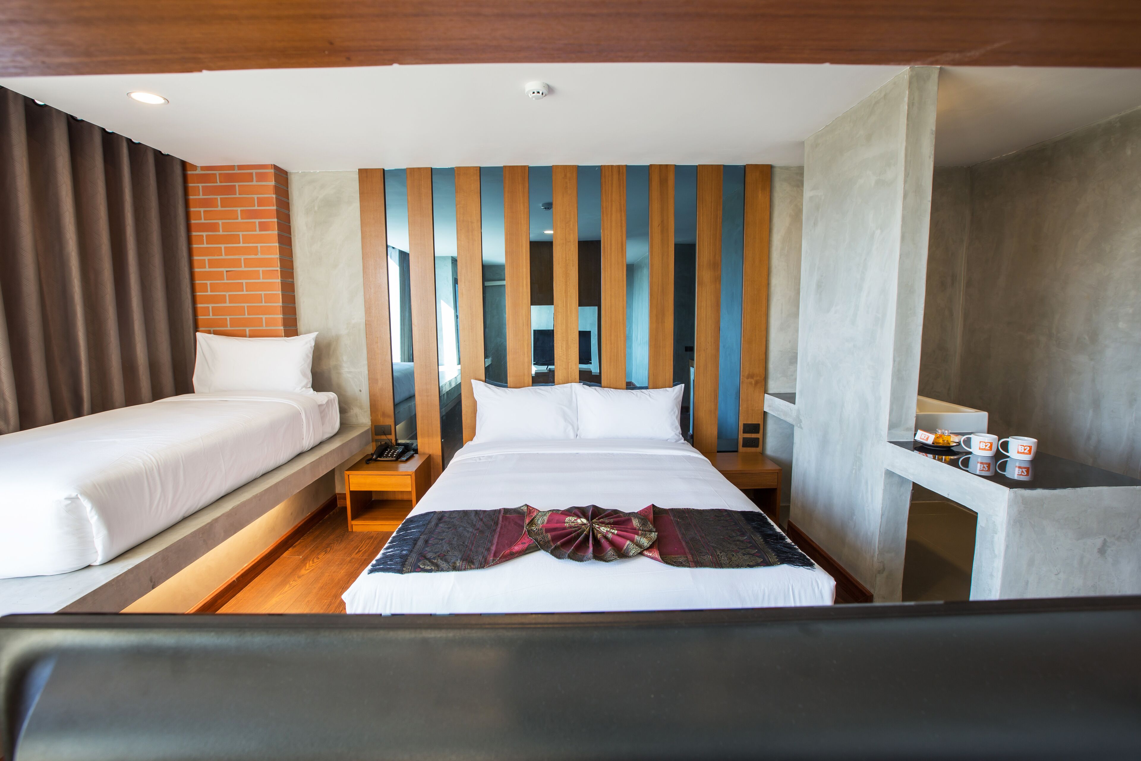 A hotel room with a bed, a nightstand, a small table with two cups, and a brick wall.
