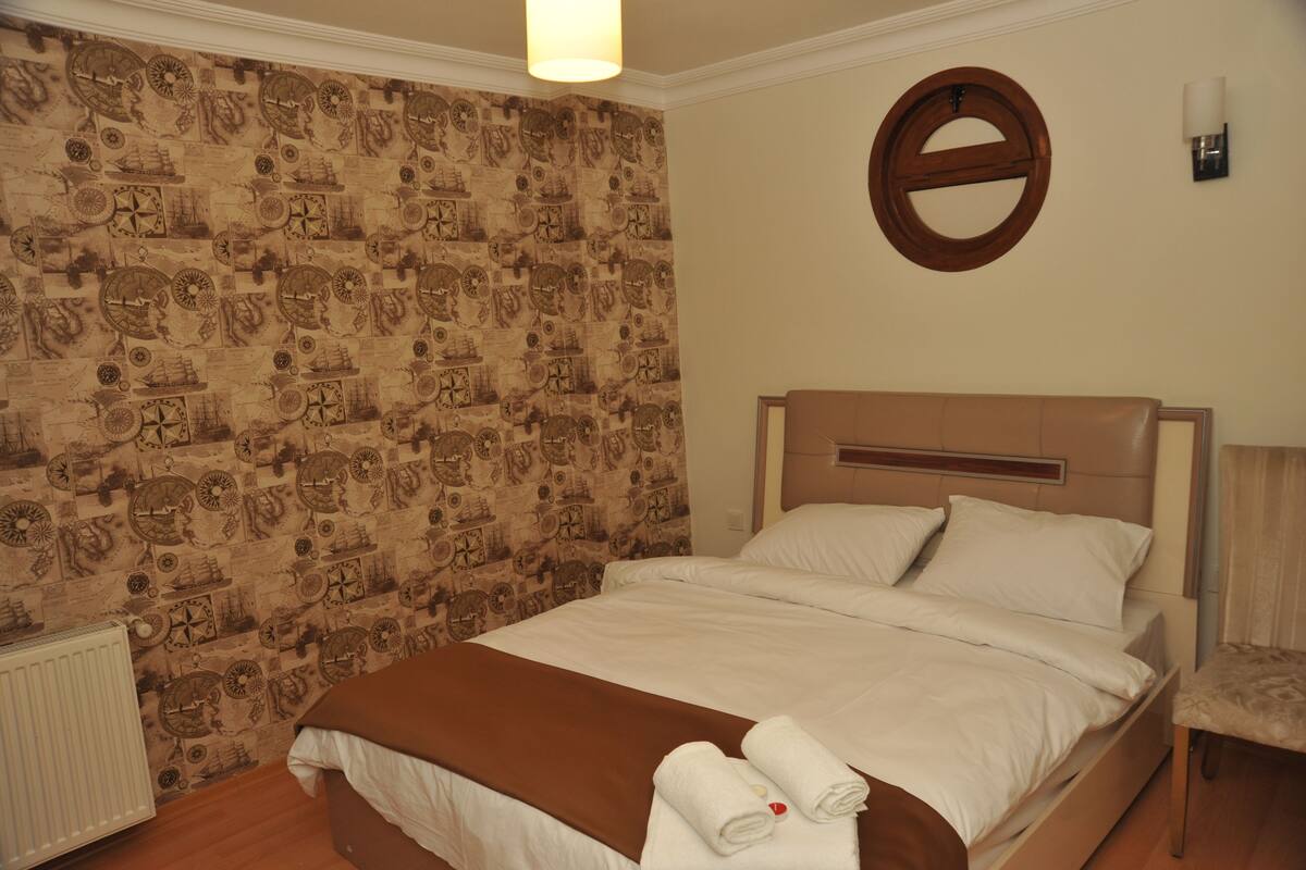 A neatly made bed with a brown blanket, a round mirror on the wall, and a patterned wallpaper.