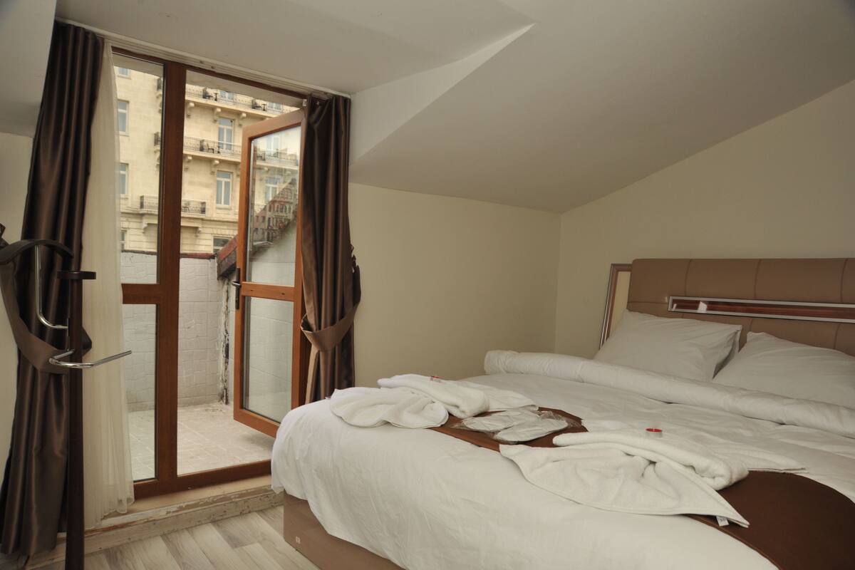 A hotel room with a bed, a window with curtains, and a view of buildings outside.