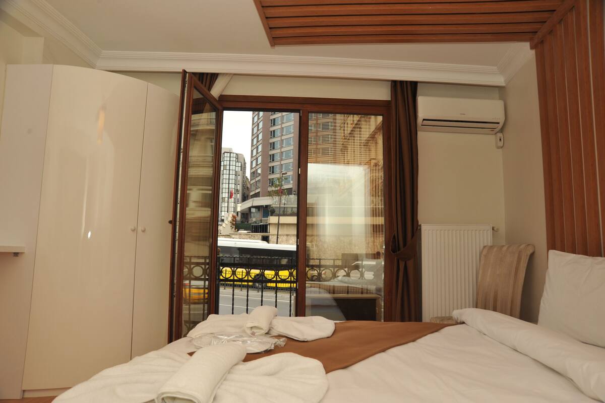 A hotel room with a bed, a wardrobe, a window with a view of buildings, and an air conditioning unit.