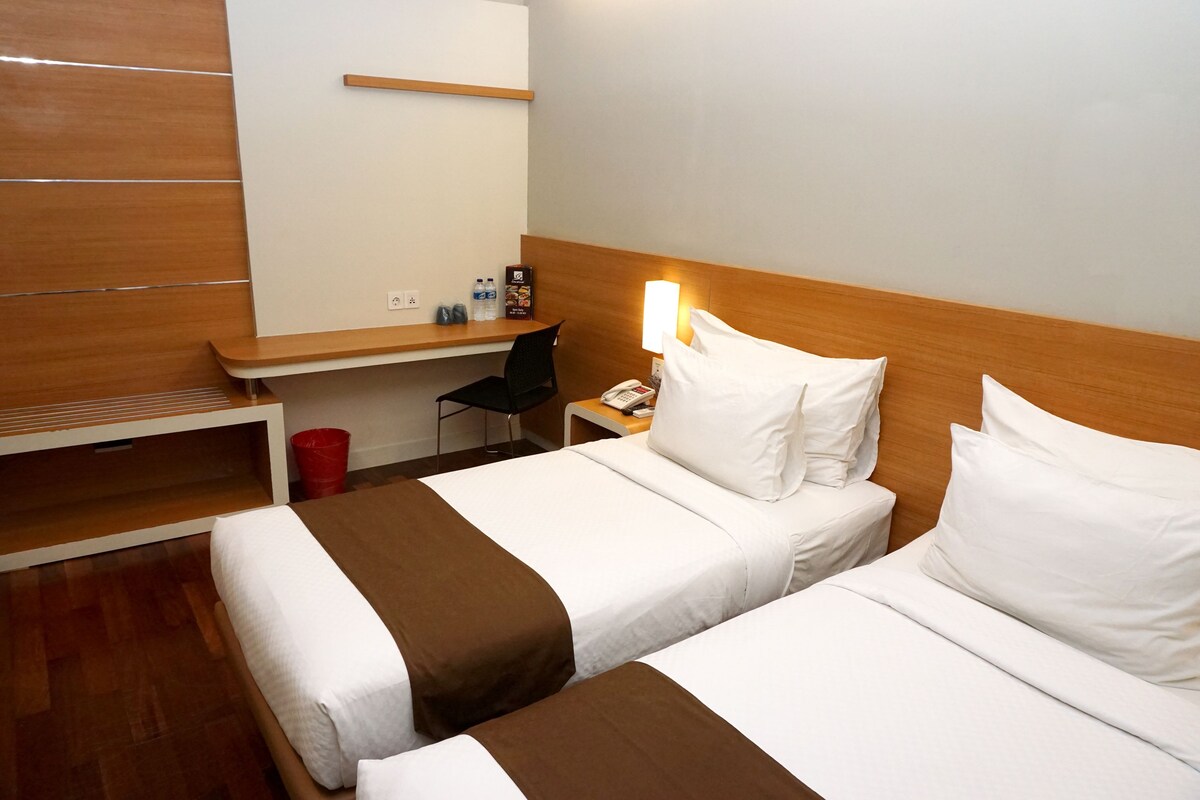 A hotel room with two beds, a desk, a chair, and a nightstand with a lamp.