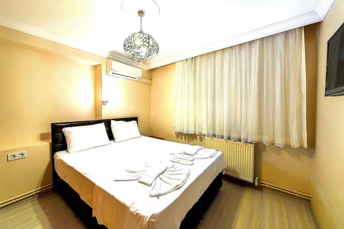 A hotel room with a bed, a radiator, a window with curtains, and a hanging light fixture.