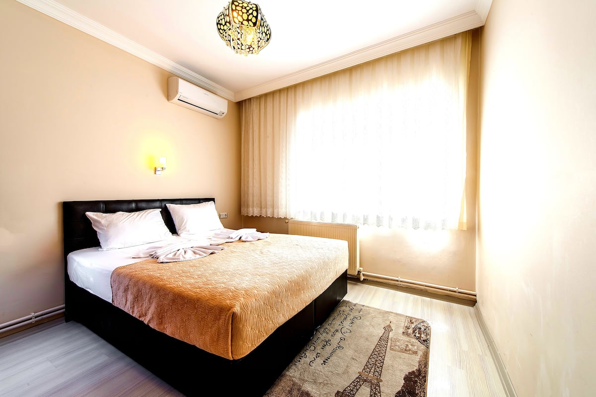 A neatly made bed with a patterned bedspread, a bedside lamp, and a view of a window with curtains.