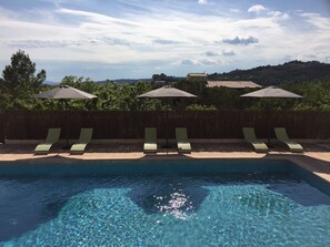 Una piscina con agua cristalina, rodeada de sillas de descanso y sombrillas, con vistas a una colina y árboles al fondo.
