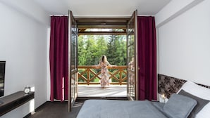 A bedroom with a large bed, a wooden headboard, a nightstand with a lamp, and a view of a balcony with a person standing outside. - Penzion Sileo (Pribylina)