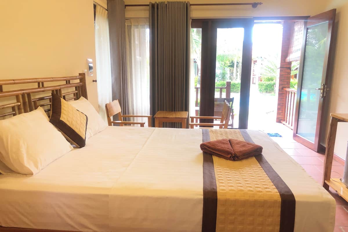 A hotel room with a large bed, wooden headboard, and a view of a garden through an open door.