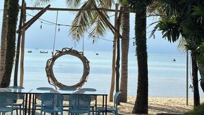 Un área de estar junto a la playa con mesas y sillas, un adorno decorativo y vistas al mar con barcos al fondo.