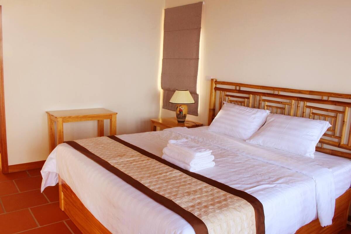 A neatly made bed with white linens and a brown bedspread, a wooden nightstand with a lamp, and a small wooden table beside the bed.
