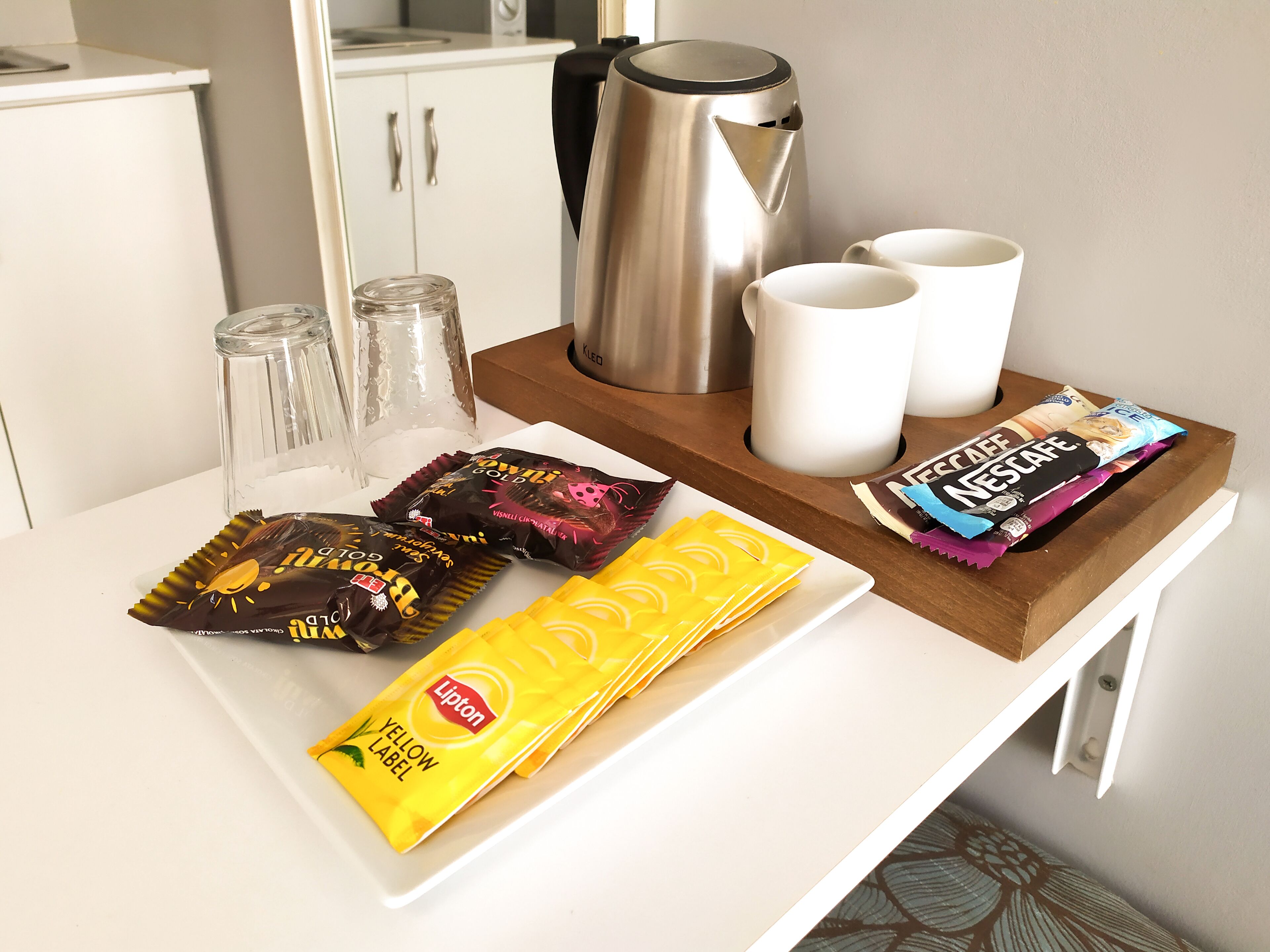A kitchen counter with a coffee maker, two mugs, a kettle, and various snack packets.