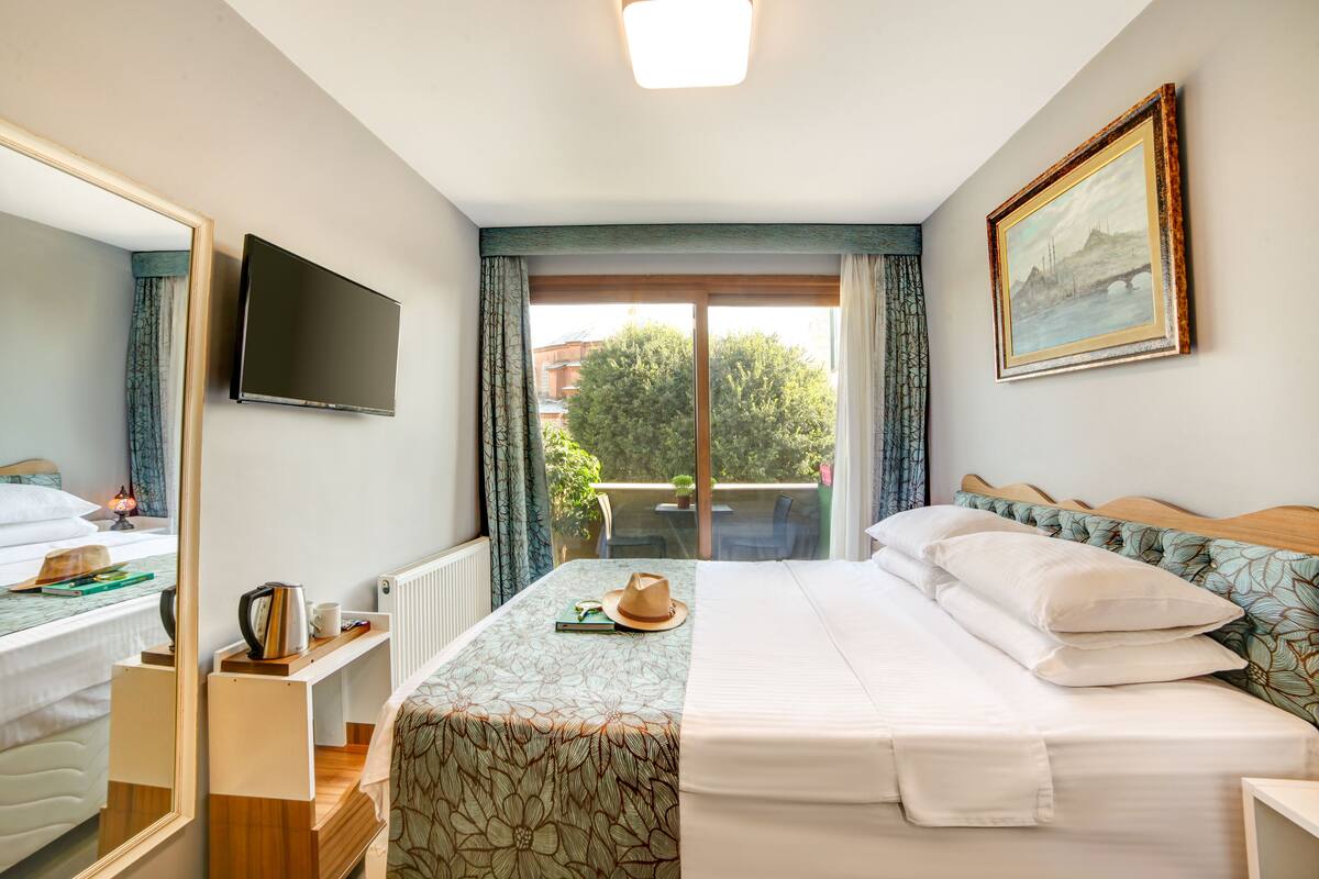 A hotel room with a bed, a TV, a window with curtains, a painting, and a view of trees and buildings outside.