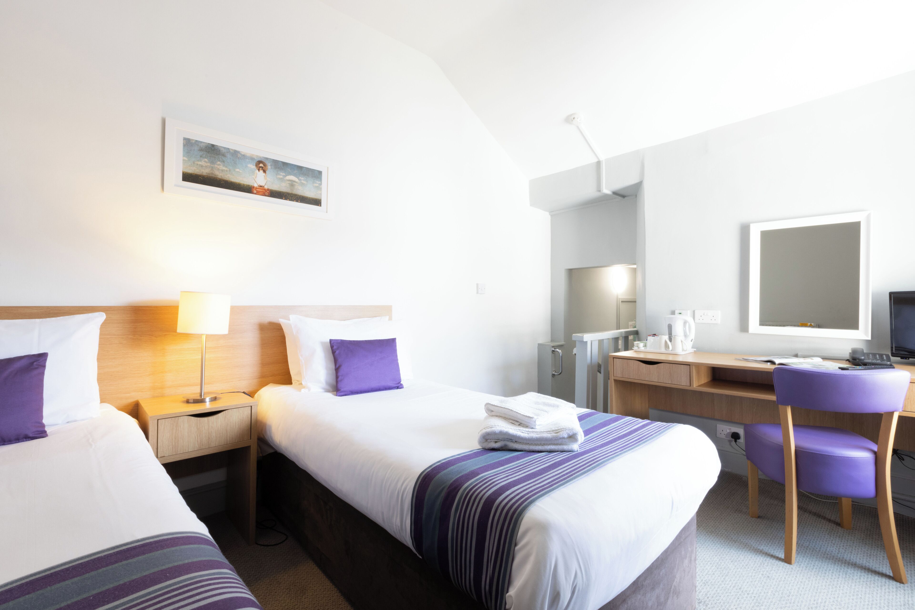 A hotel room with two beds, a desk, a chair, a desk lamp, a coffee maker, and a large mirror.