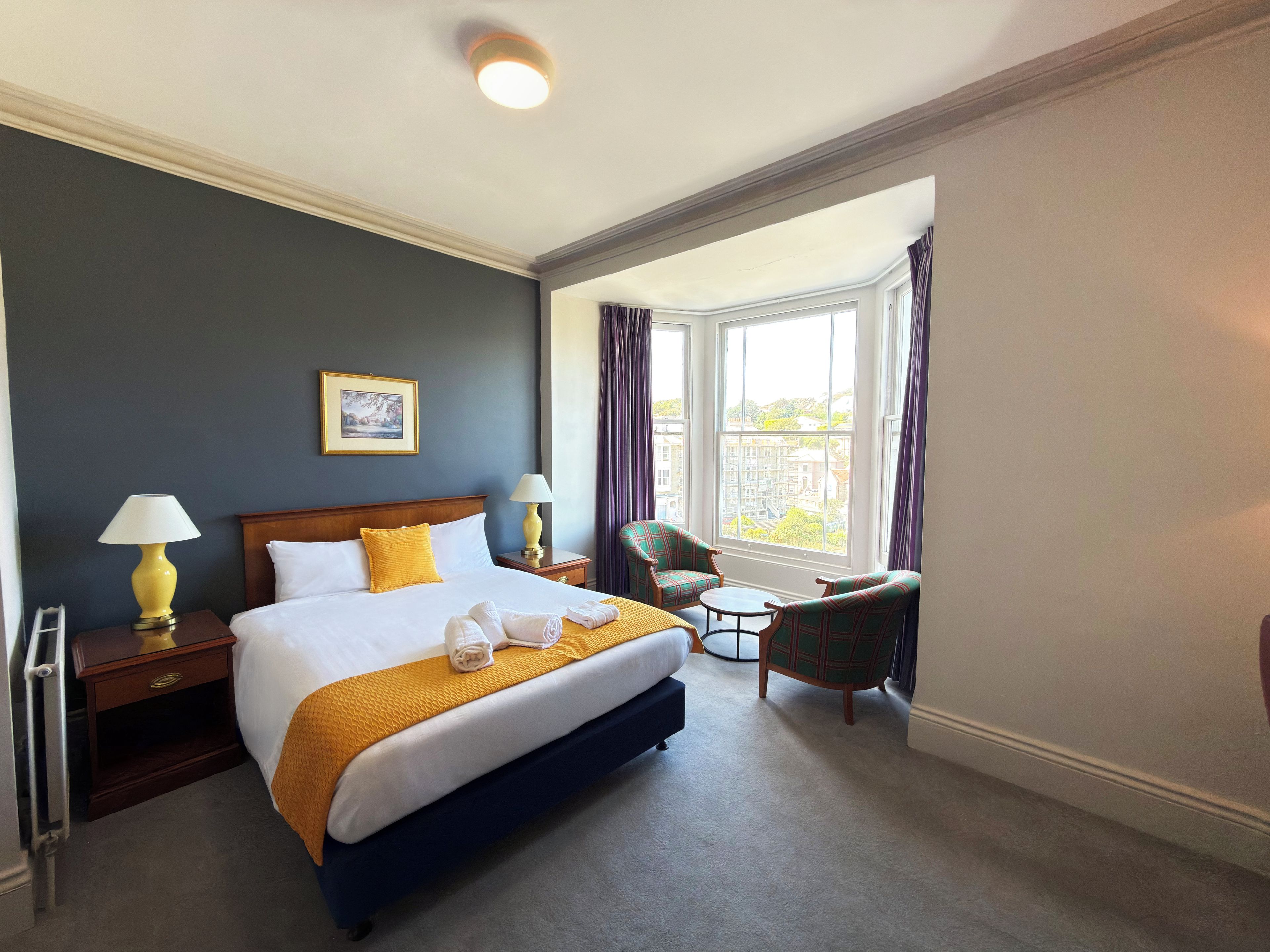 A hotel room with a bed, two bedside tables, a chair, a small table, and a window with curtains.
