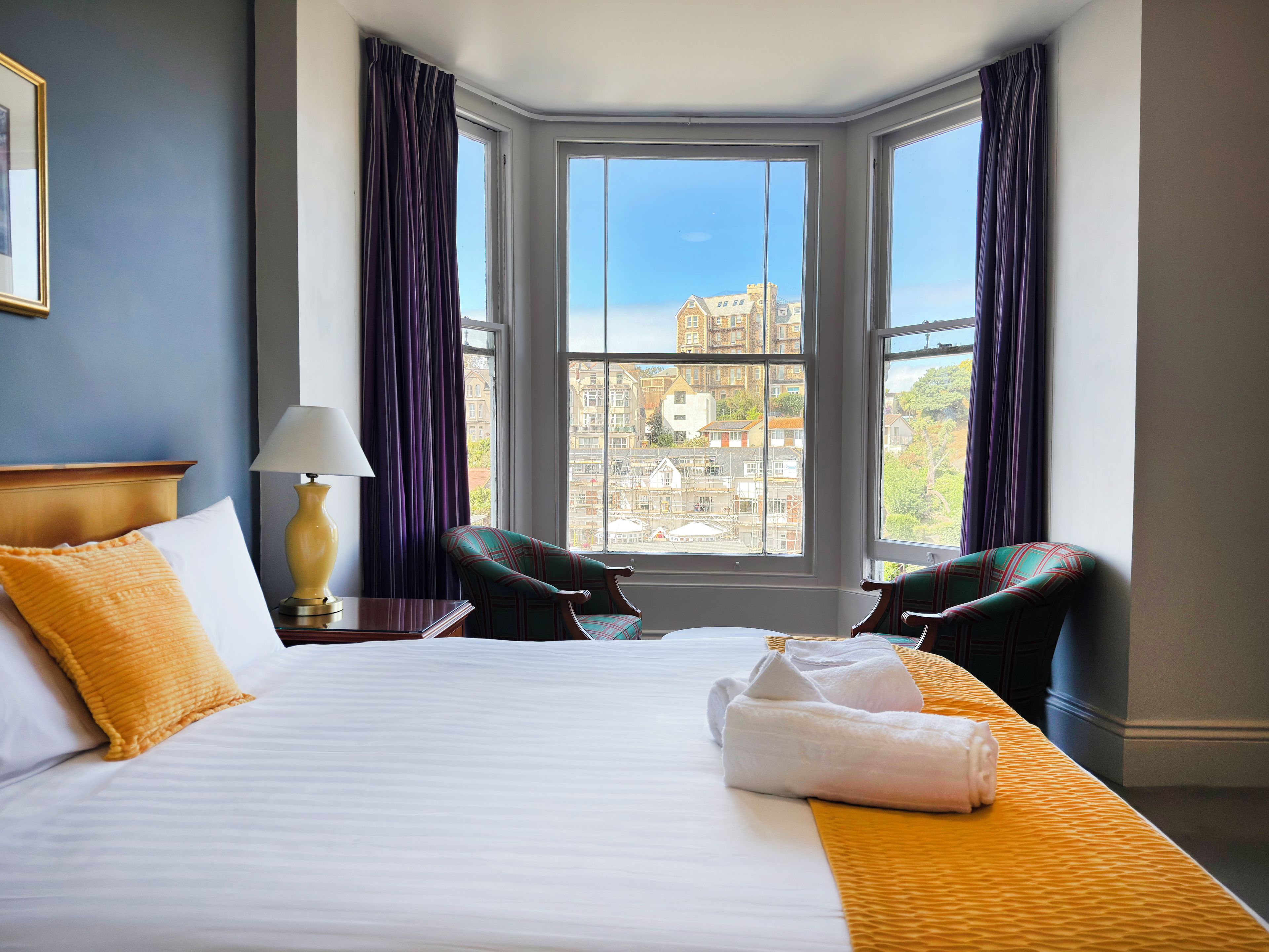 A hotel room with a large bed, two chairs, a lamp, and a view of buildings through the window.
