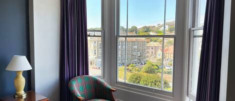 A hotel room with a large bay window, a plaid armchair, a small round table, and a bedside lamp.