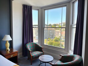 A hotel room with a large bay window, a plaid armchair, a small round table, and a bedside lamp. - The Dilkhusa Grand Hotel By Compass Hospitality (Ilfracombe)