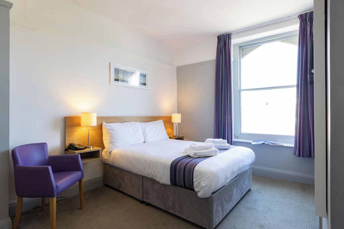 Een hotelkamer met een bed, een stoel, een nachtkastje en een raam met gordijnen.