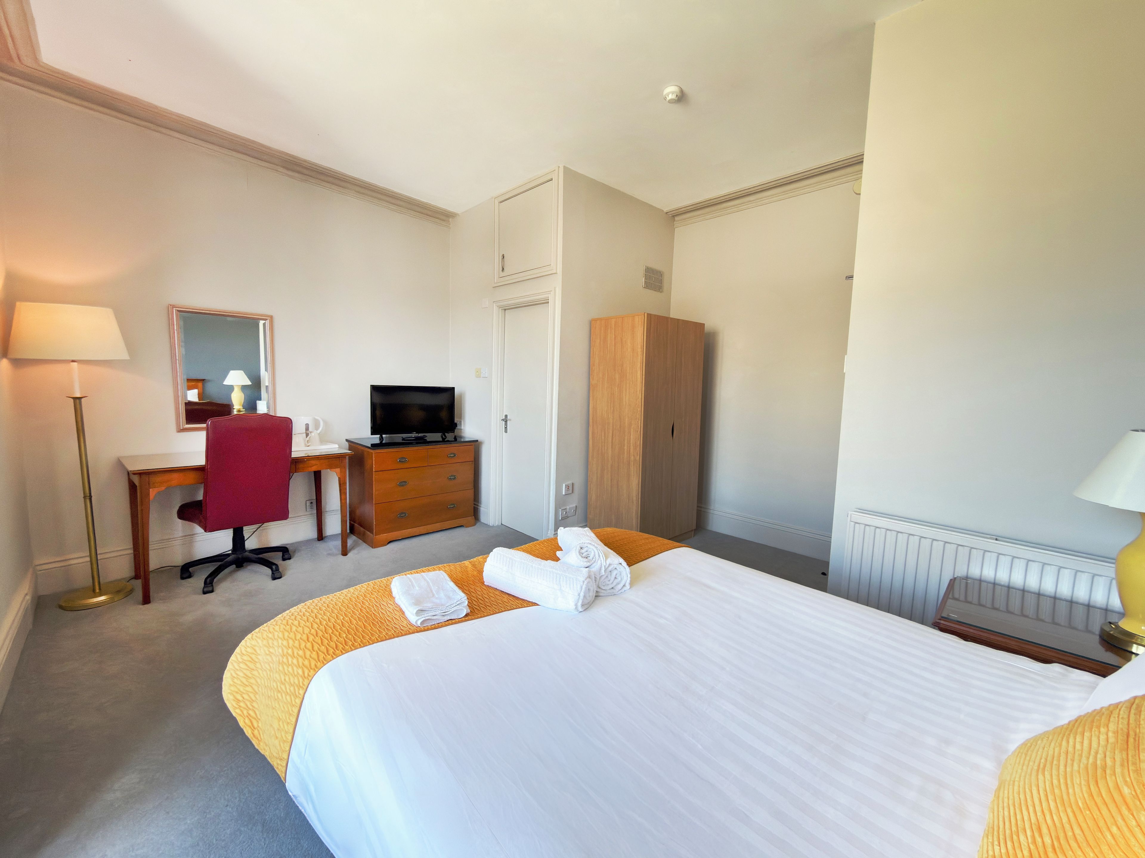 A hotel room with a bed, a desk with a television, a wardrobe, and a lamp.