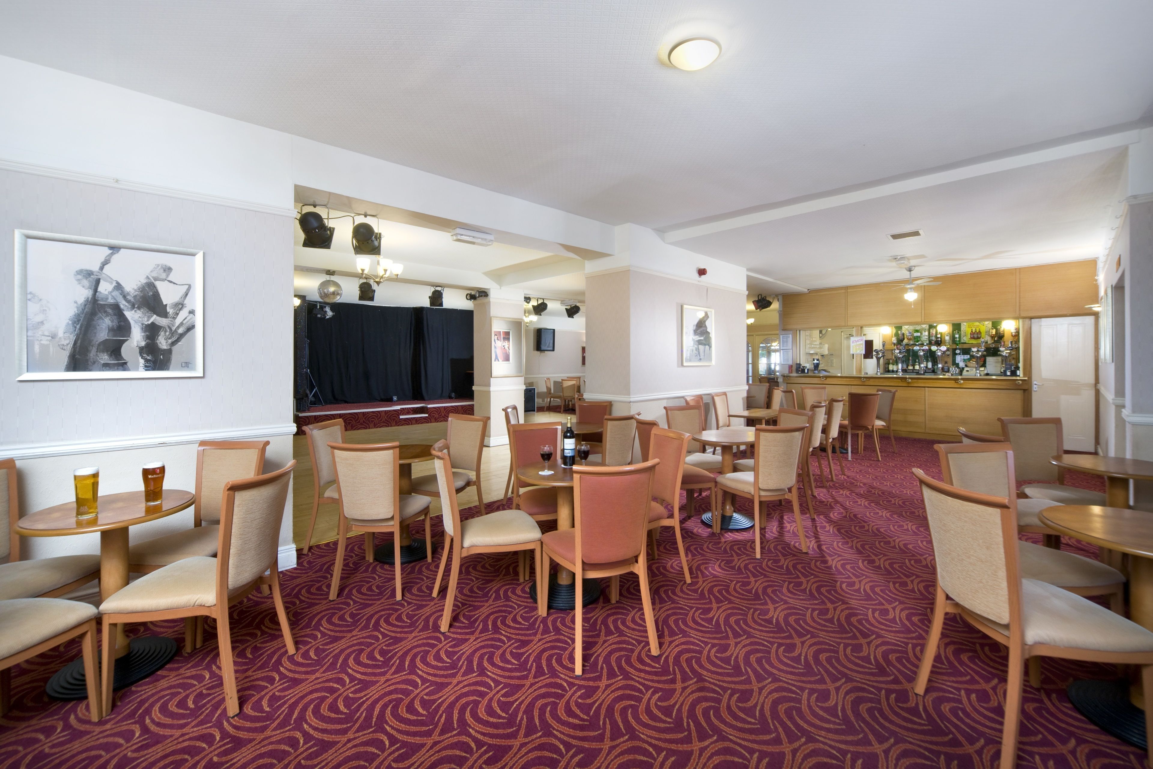 A hotel bar/lounge area with multiple seating arrangements, including round tables and chairs, and a bar counter with various bottles.