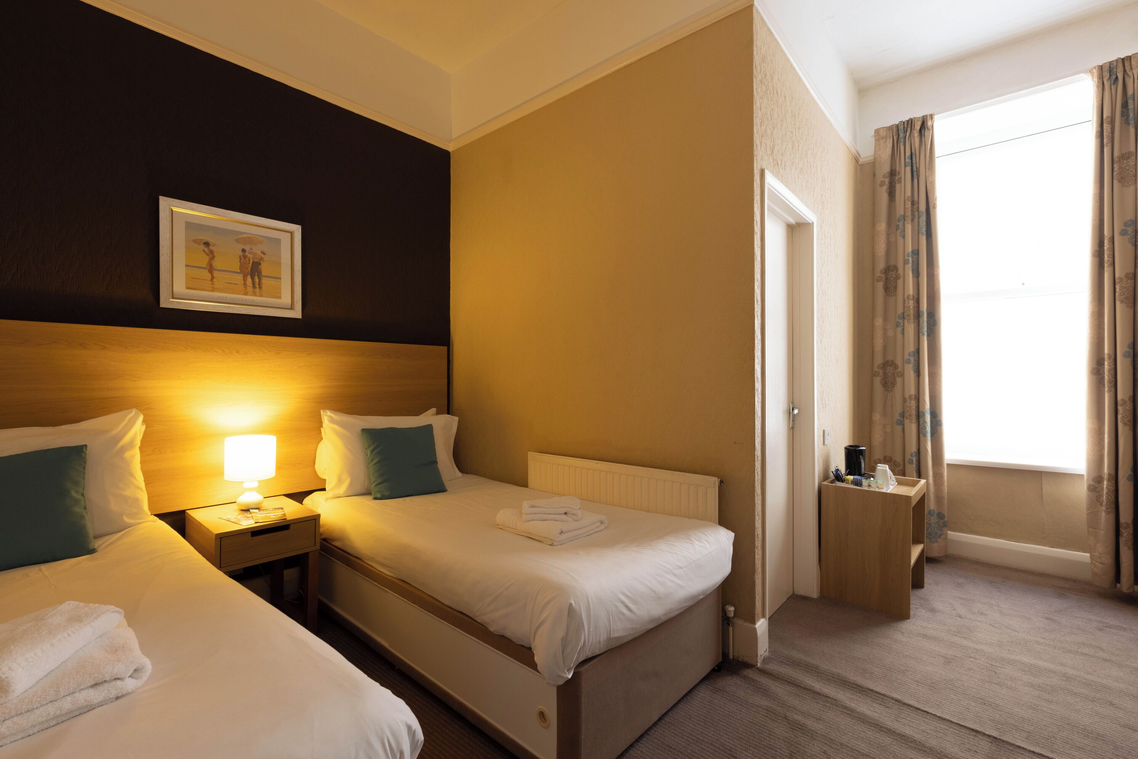 Habitación de hotel con dos camas, una mesita de noche con lámpara, un cuadro en la pared y una ventana con cortinas.