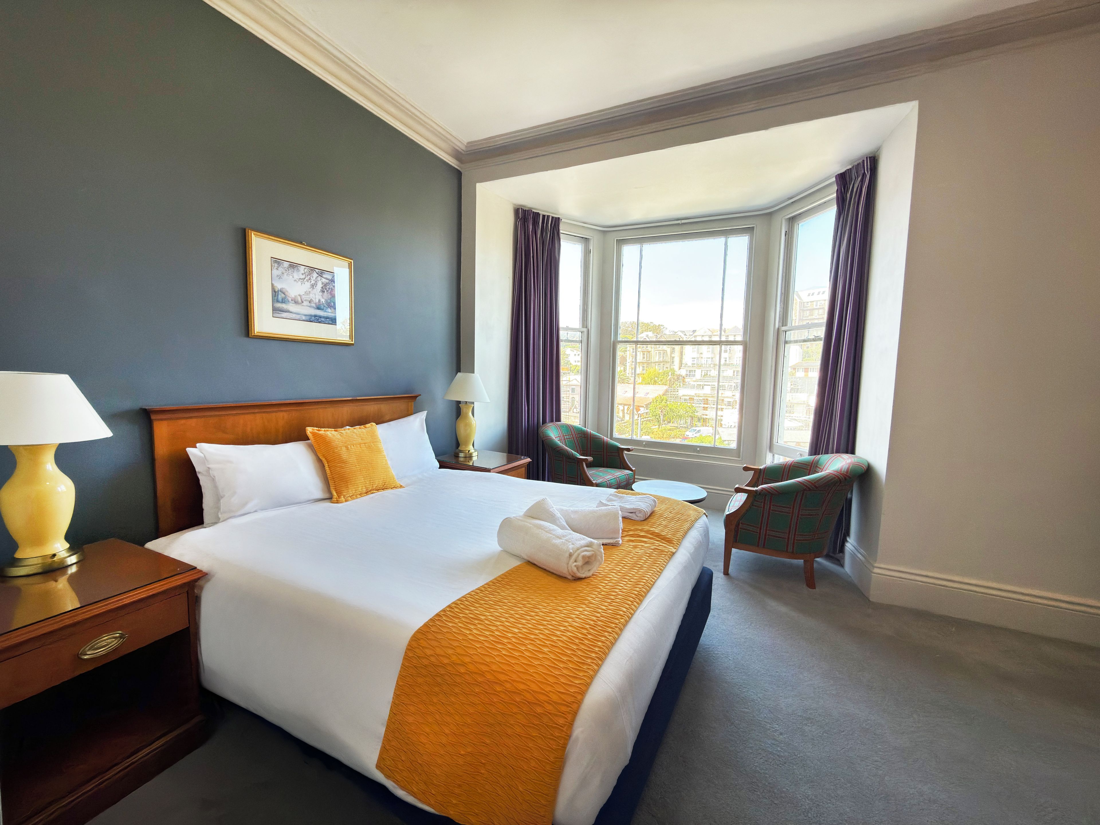 A hotel room with a large bed, a desk, two chairs, a window with curtains, and a painting on the wall.