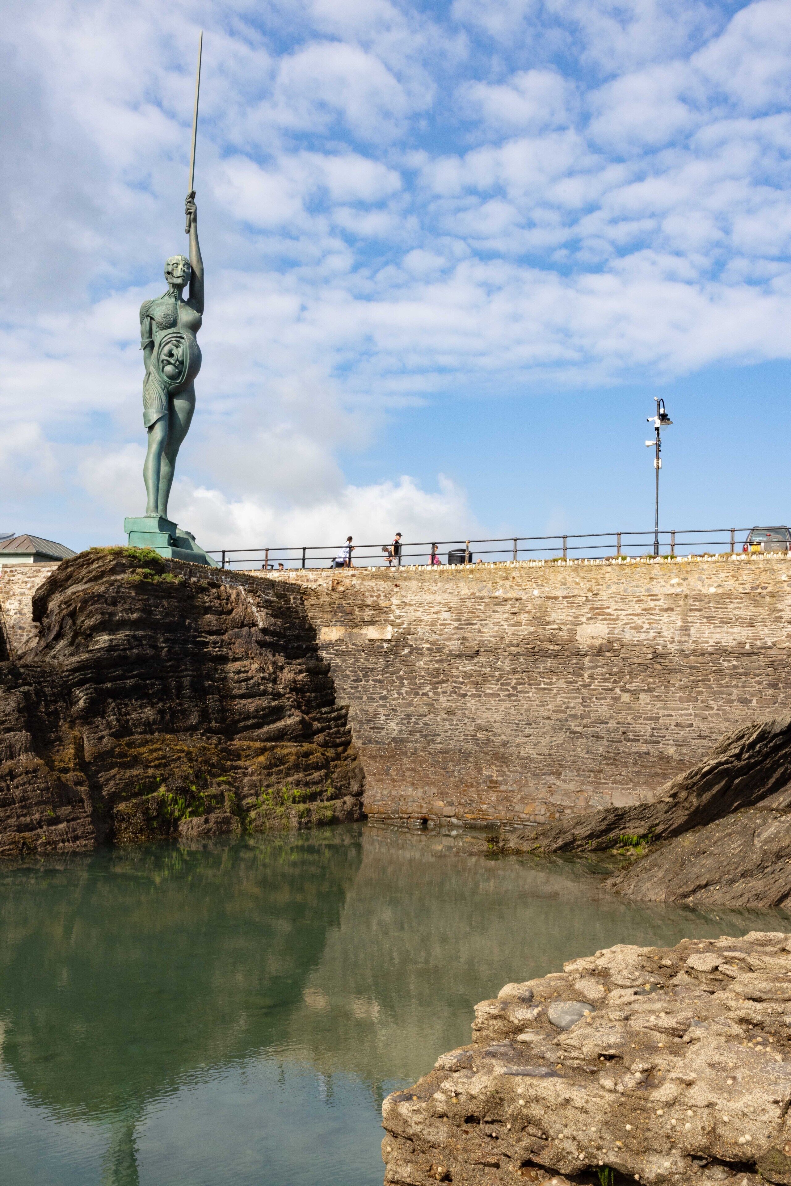 A statue on a rock by a body of water, with a stone wall and a railing in the background.