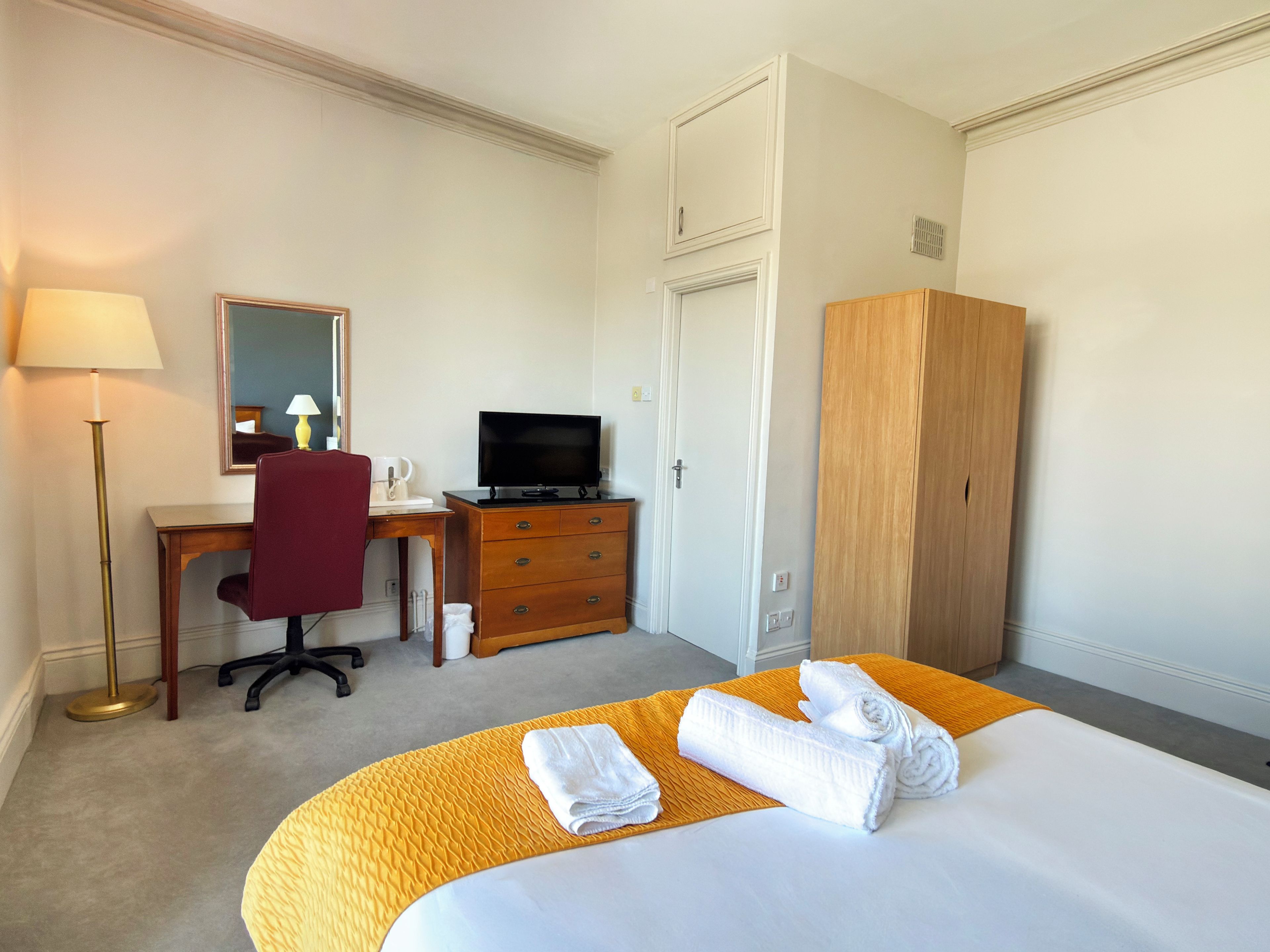 A hotel room with a bed, a desk with a television, a wardrobe, and a lamp.