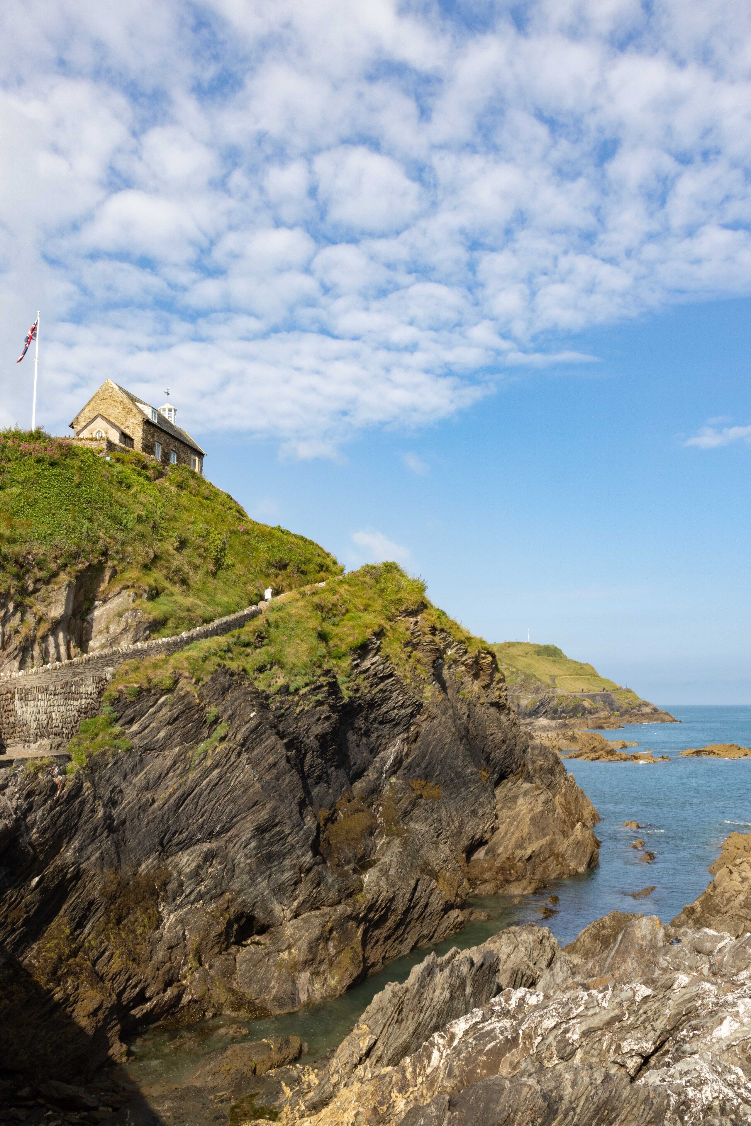 A coastal building with a stone facade, situated on a grassy hill overlooking the sea.