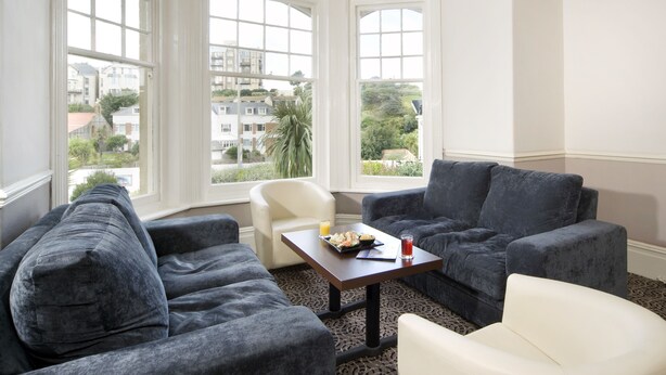 Een kamer met een donkergrijze bank, een witte fauteuil en een bijzettafeltje met een drankje en een bord eten. Grote ramen bieden uitzicht op gebouwen en groen buiten.