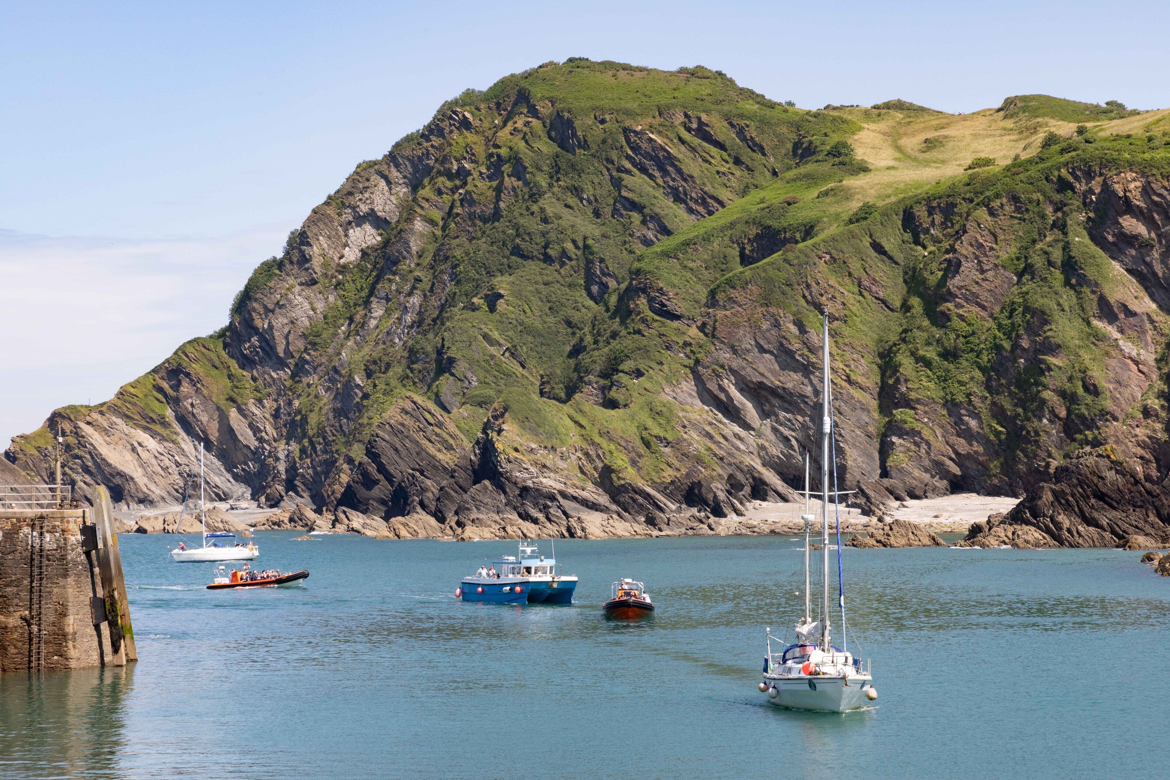 A coastal scene with boats on calm water, a rocky cliff, and a lighthouse.
