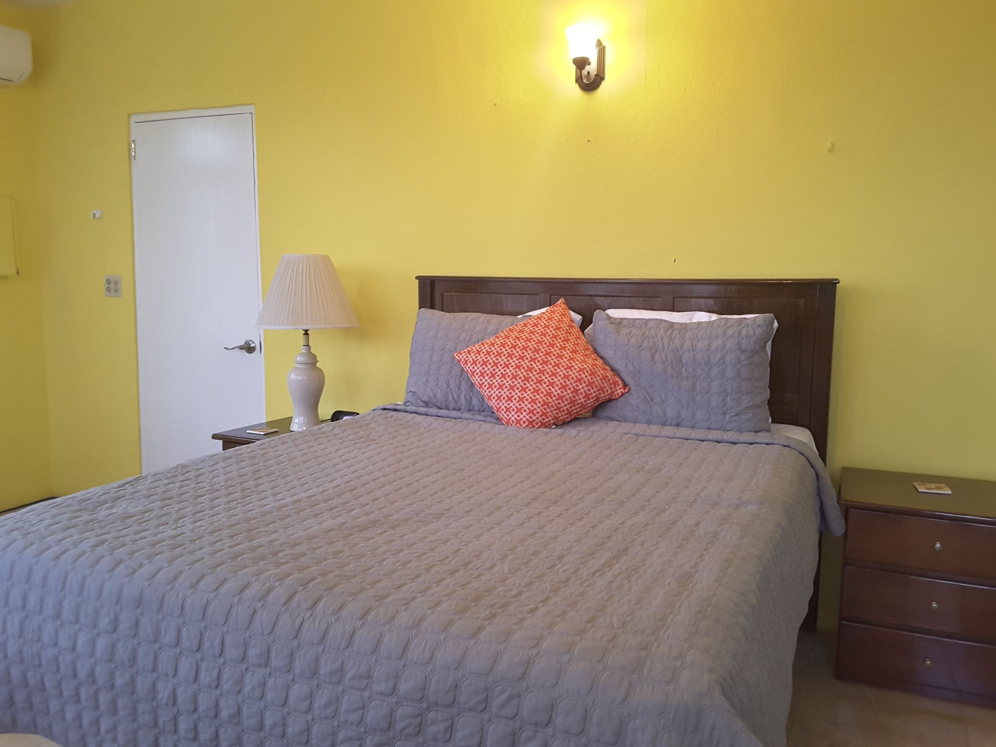 A bed with a grey quilt, a wooden headboard, and two pillows (one red and one grey).