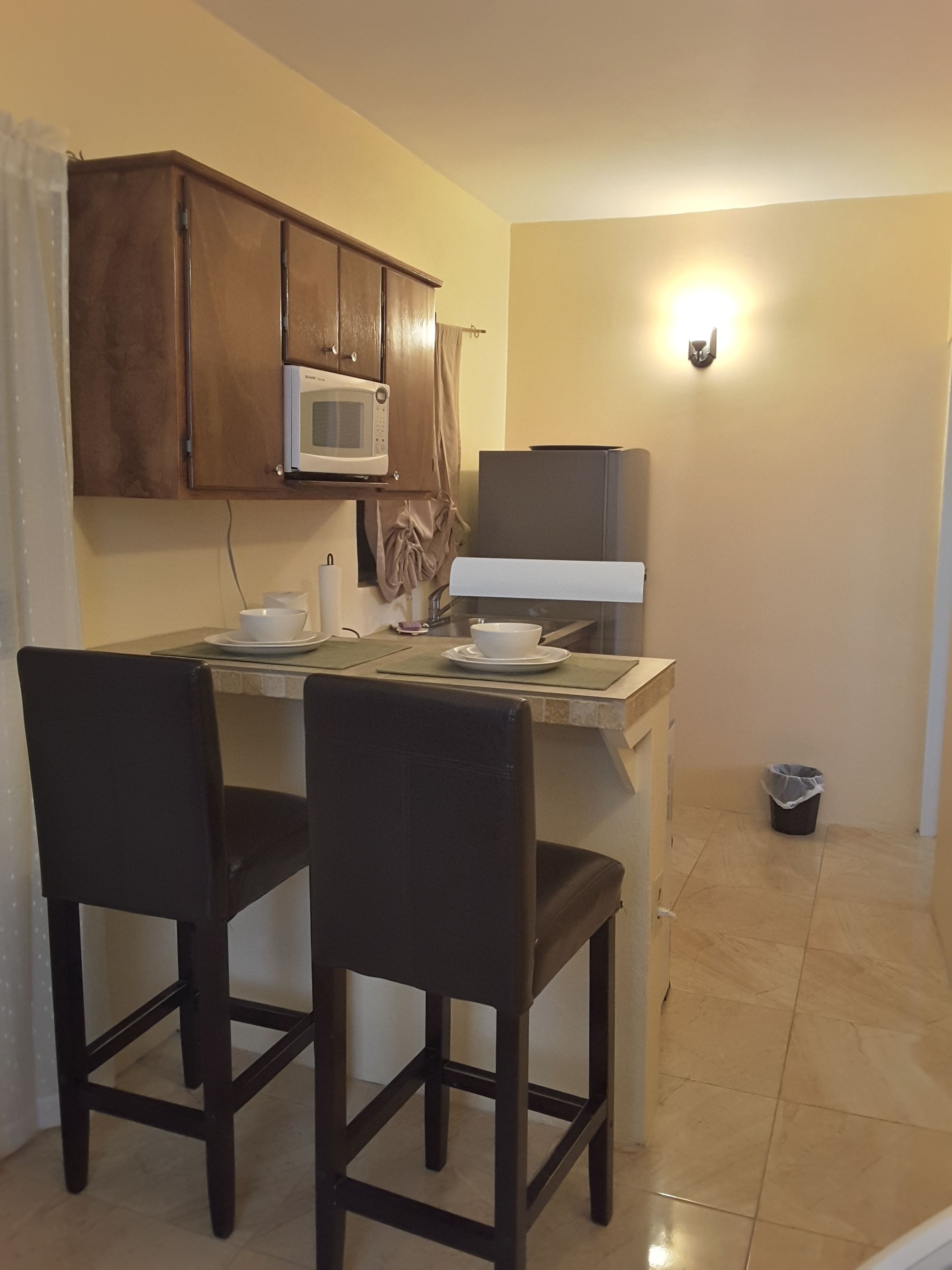 Cocina compacta con microondas, lavamanos y refrigerador pequeño. Hay dos sillas y una mesa.