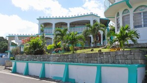 A two-story building with a white and teal color scheme, featuring balconies and a stone retaining wall. - The Apartments at Jolly Castle (Jolly Harbour)
