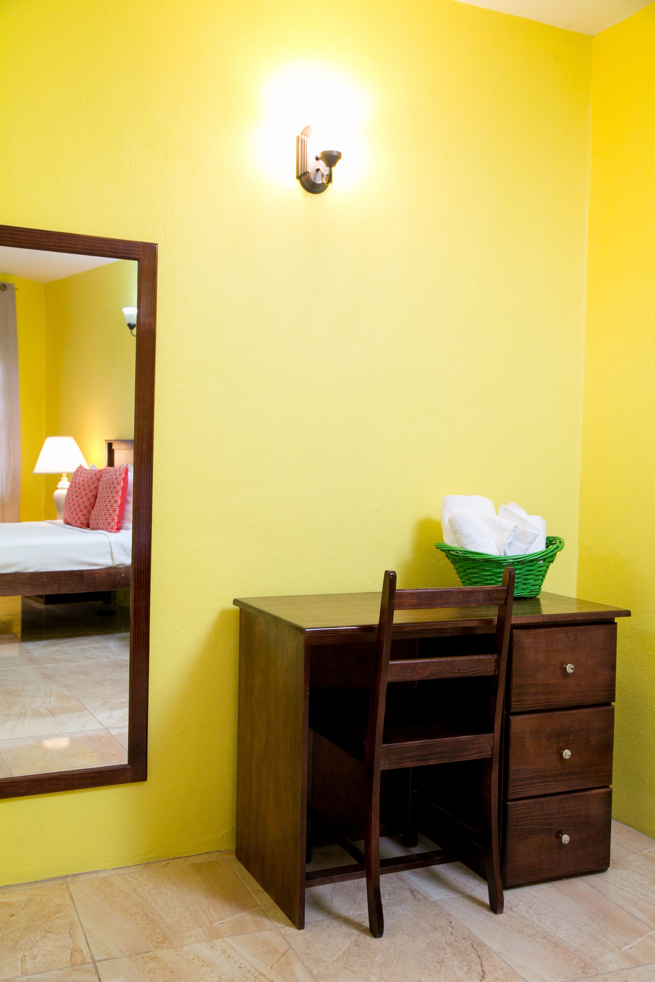 A room with a wooden desk, a chair, a green basket with towels, a mirror, and a wall-mounted light fixture.