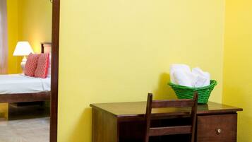 A room with a wooden desk, a chair, a green basket with towels, a mirror, and a wall-mounted light fixture.