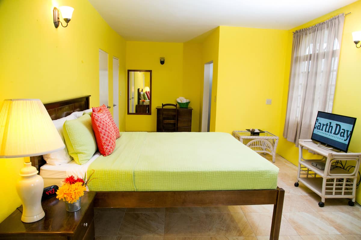 A bright yellow room with a bed, a nightstand with a lamp, a TV on a stand, and a mirror on the wall.