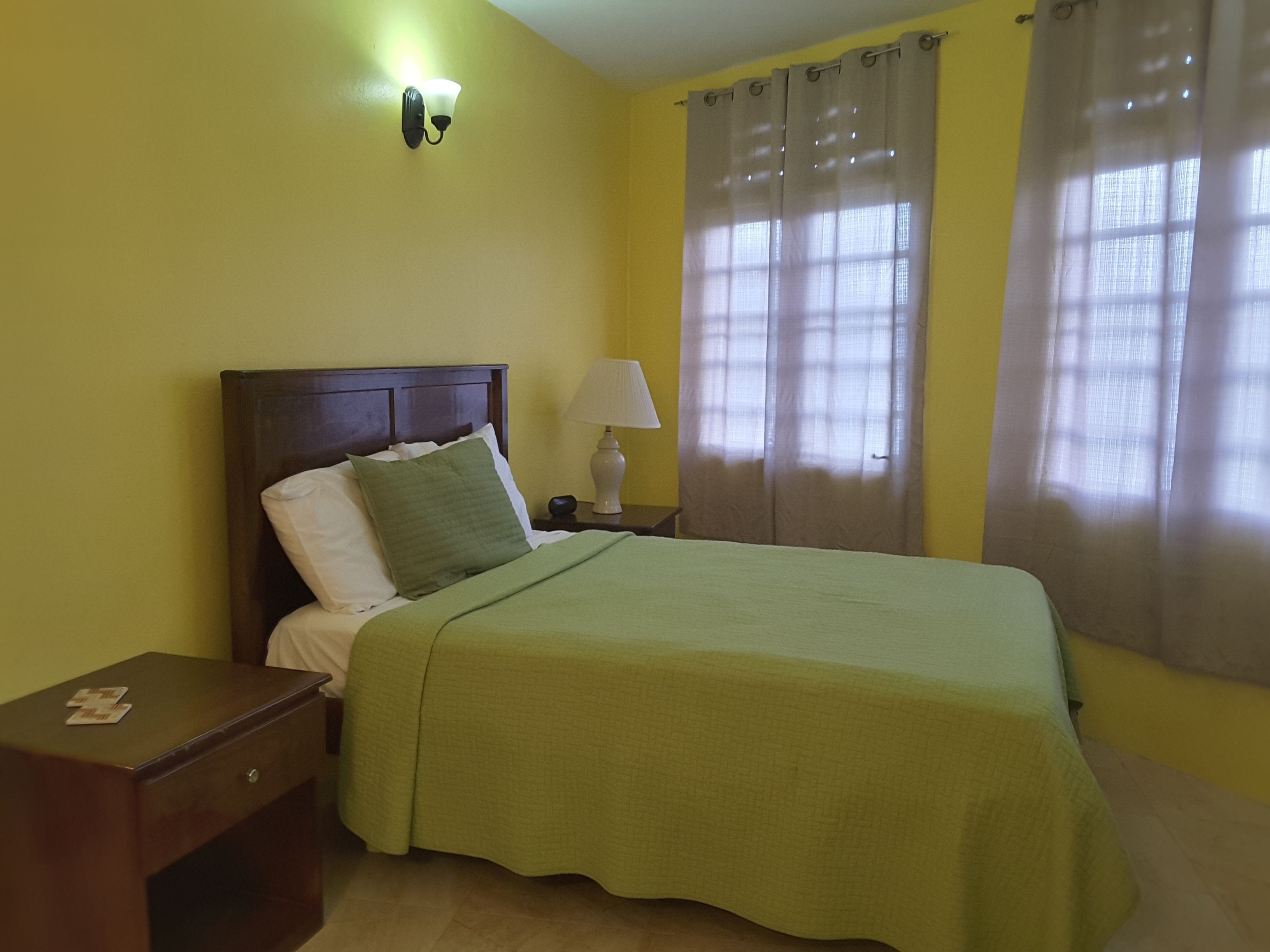 A neatly made bed with a green bedspread, a wooden nightstand with a lamp, and a window with sheer curtains.