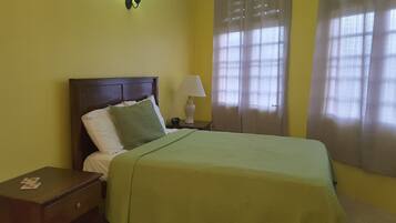 A neatly made bed with a green bedspread, a wooden nightstand with a lamp, and a window with sheer curtains.