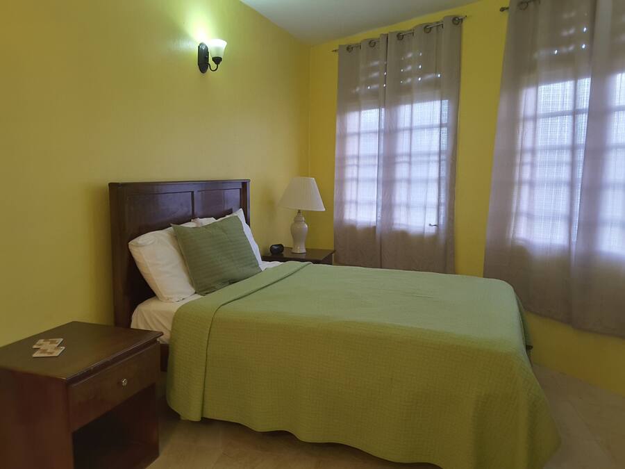 A neatly made bed with a green bedspread, a wooden nightstand with a lamp, and a window with sheer curtains.