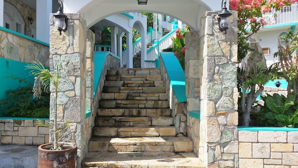 A stone staircase leading to a white building with turquoise accents, surrounded by potted plants and flowers. - The Apartments at Jolly Castle (Jolly Harbour)